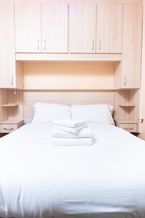 A neatly made bed with white linens and a stack of folded towels, a wooden headboard, and light-colored wooden cabinets above the bed.