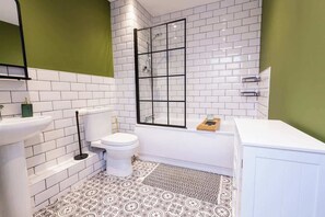 A bathroom with white tiled walls, a patterned floor, a toilet, a bathtub, and a green accent wall. - Modern Retreat: 2BR Apt with Lush Greenery (Sheffield)