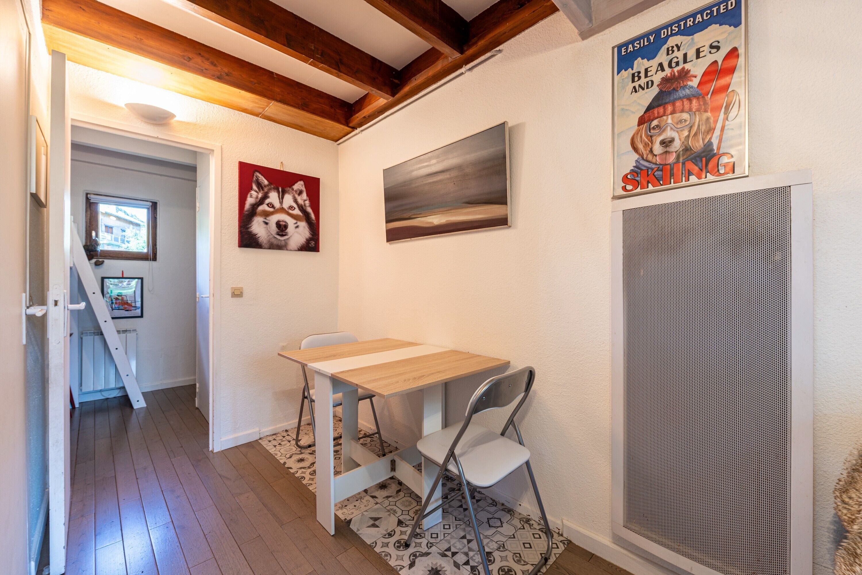 Een kamer met een houten vloer, een witte tafel met stoel, een schilderij van een hond, een raam en een deur.