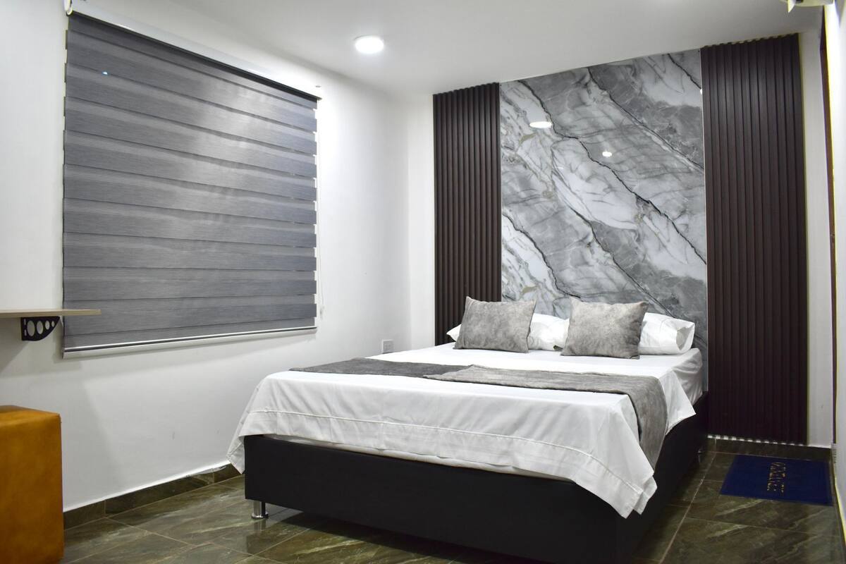 Une chambre à coucher moderne avec un lit, une fenêtre avec des stores, un mur en marbre et une tête de lit de couleur sombre.