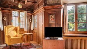 A room with a wooden interior, a yellow armchair, a television on a wooden cabinet, and a large window with a view of greenery. - Room 'Hirschen Fürstenzimmer' with Mountain View, Shared Terrace and Wi-Fi (Sarentino)