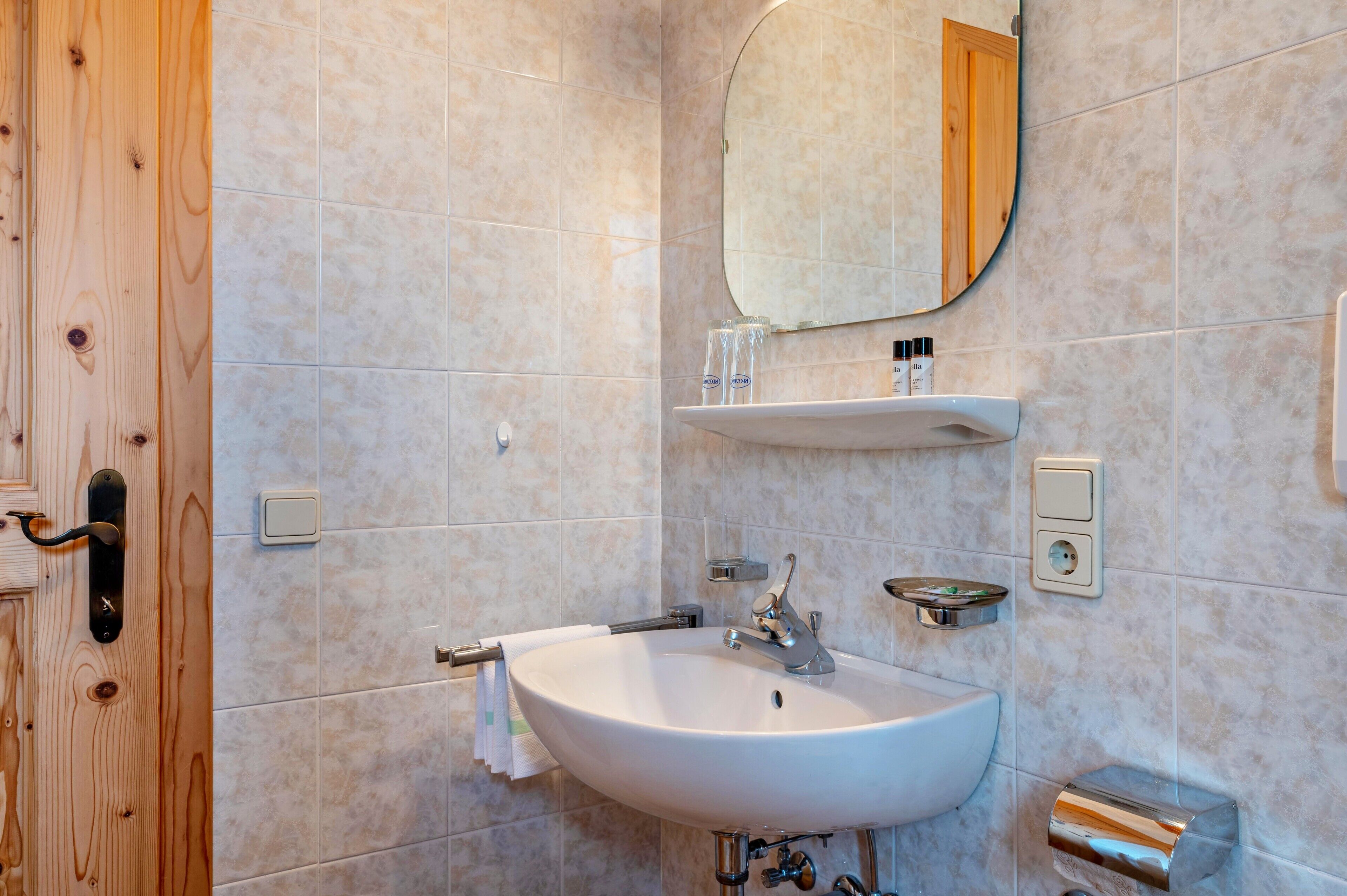 Ein Badezimmer mit Holztür, Waschbecken in Weiß, einem Spiegel und einem Ablagebereich für Toilettenartikel.