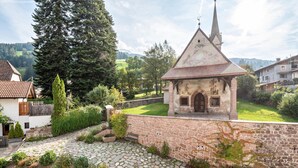 A stone church with a pointed roof, surrounded by a stone wall and greenery, with a cobblestone path leading to it.