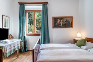 A bedroom with a bed, a table with a blanket, a painting, and a window with curtains.