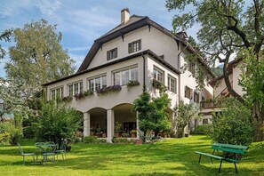 A large, two-story building with a covered patio area, surrounded by lush greenery and a well-maintained lawn.