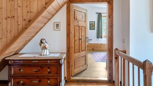 Eine Holztreppe führt zu einem Raum mit einer Holztür, einer kleinen Statue auf einer Kommode und einem Fenster mit Vorhängen.
