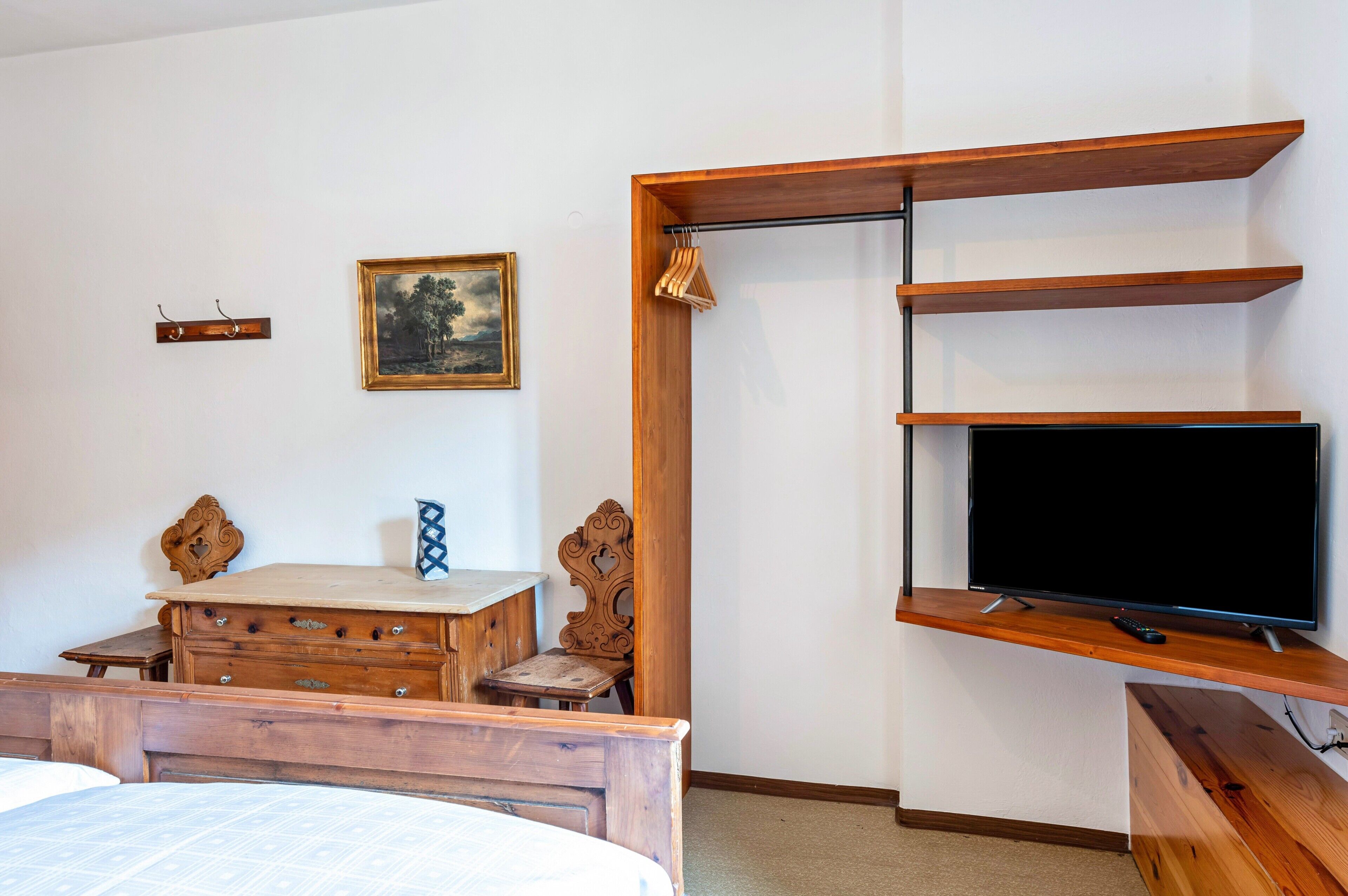 A bedroom with a bed, wooden dresser, desk, chair, TV mounted on a wooden shelf, and a painting on the wall.