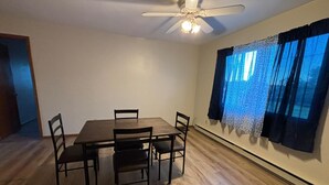 A dining room with a wooden table and chairs, a ceiling fan, and a window with patterned curtains.