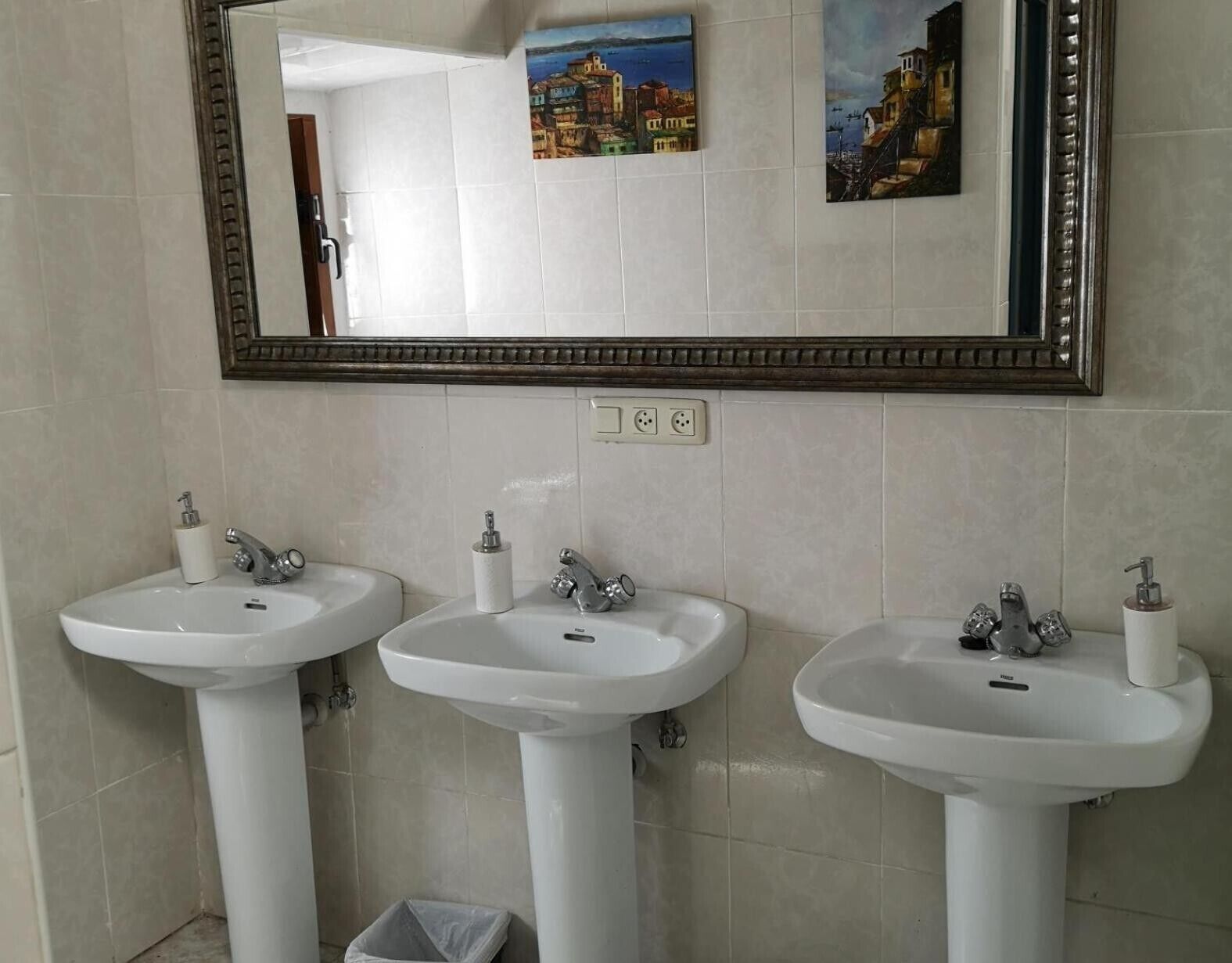 Un baño con tres lavamanos blancos, un espejo y dos cuadros colgados en la pared.