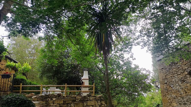Una escalera de piedra conduce a una terraza de madera con una chimenea de piedra, rodeada de exuberante follaje y flores moradas.