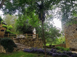 A stone staircase leading up to a wooden deck with a stone chimney, surrounded by lush greenery and purple flowers.