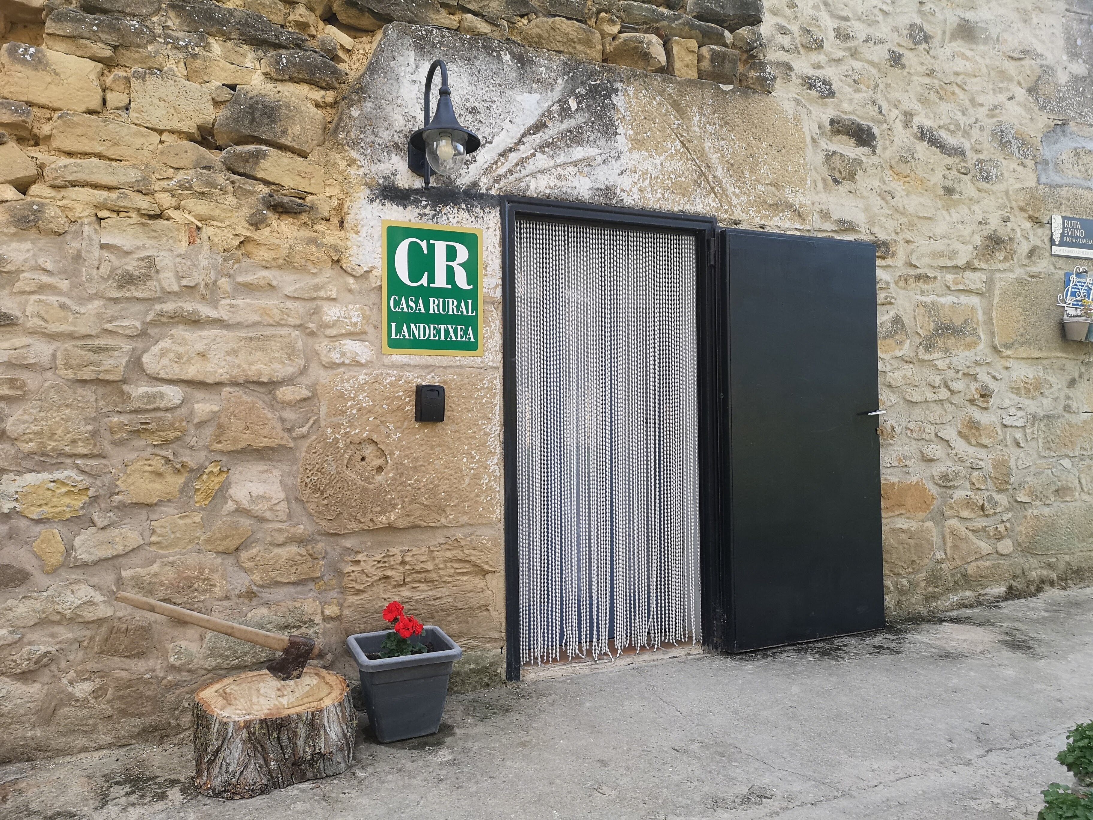 Un mur de pierres rustiques surmonté d’un portail en métal, une enseigne verte indiquant « CR CASA RURAL LANDETXEA », et une petite plante en pot.