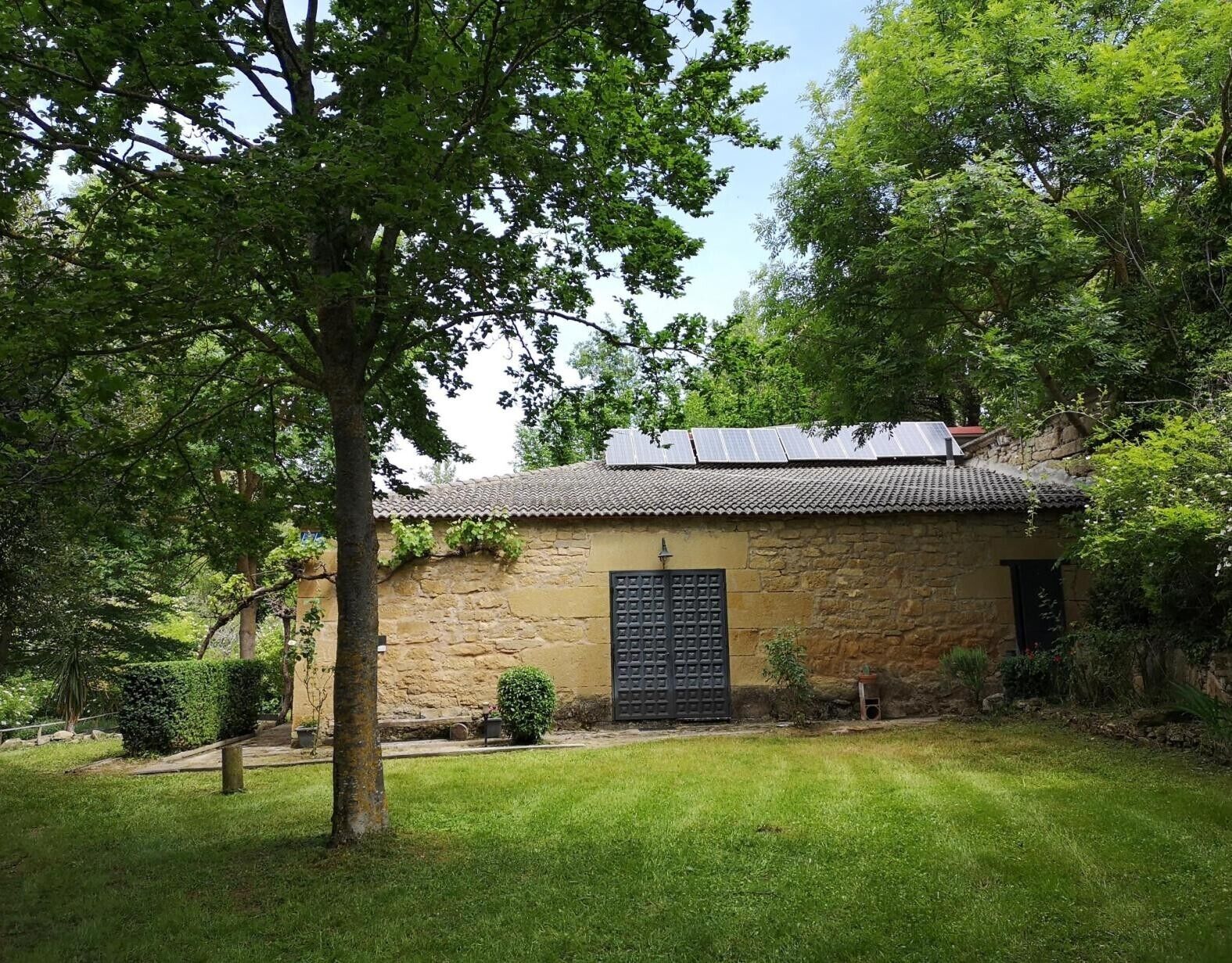 A stone building with a tiled roof and a black door, surrounded by greenery and trees.
