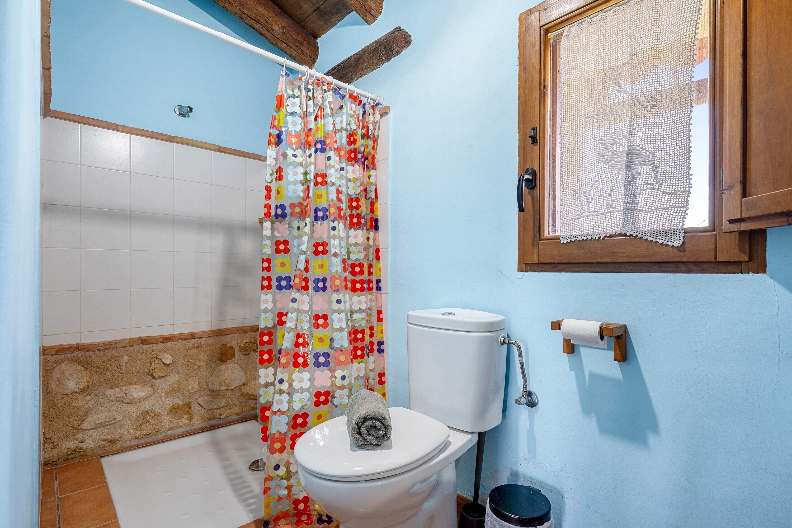 A bathroom with a toilet, a shower curtain, a window with a lace curtain, and a tiled wall.