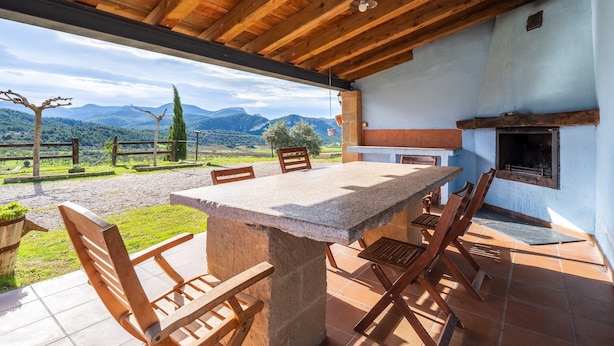An outdoor dining area with a stone table and wooden chairs, a fireplace, and a view of mountains. - Country House 'La Sala' with Mountain View, Shared Pool and Wi-Fi (Peñarroya De Tastavins)