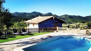A pool area with a wooden fence, a blue building, and mountains in the background. - Country House 'La Sala' with Mountain View, Shared Pool and Wi-Fi (Peñarroya De Tastavins)