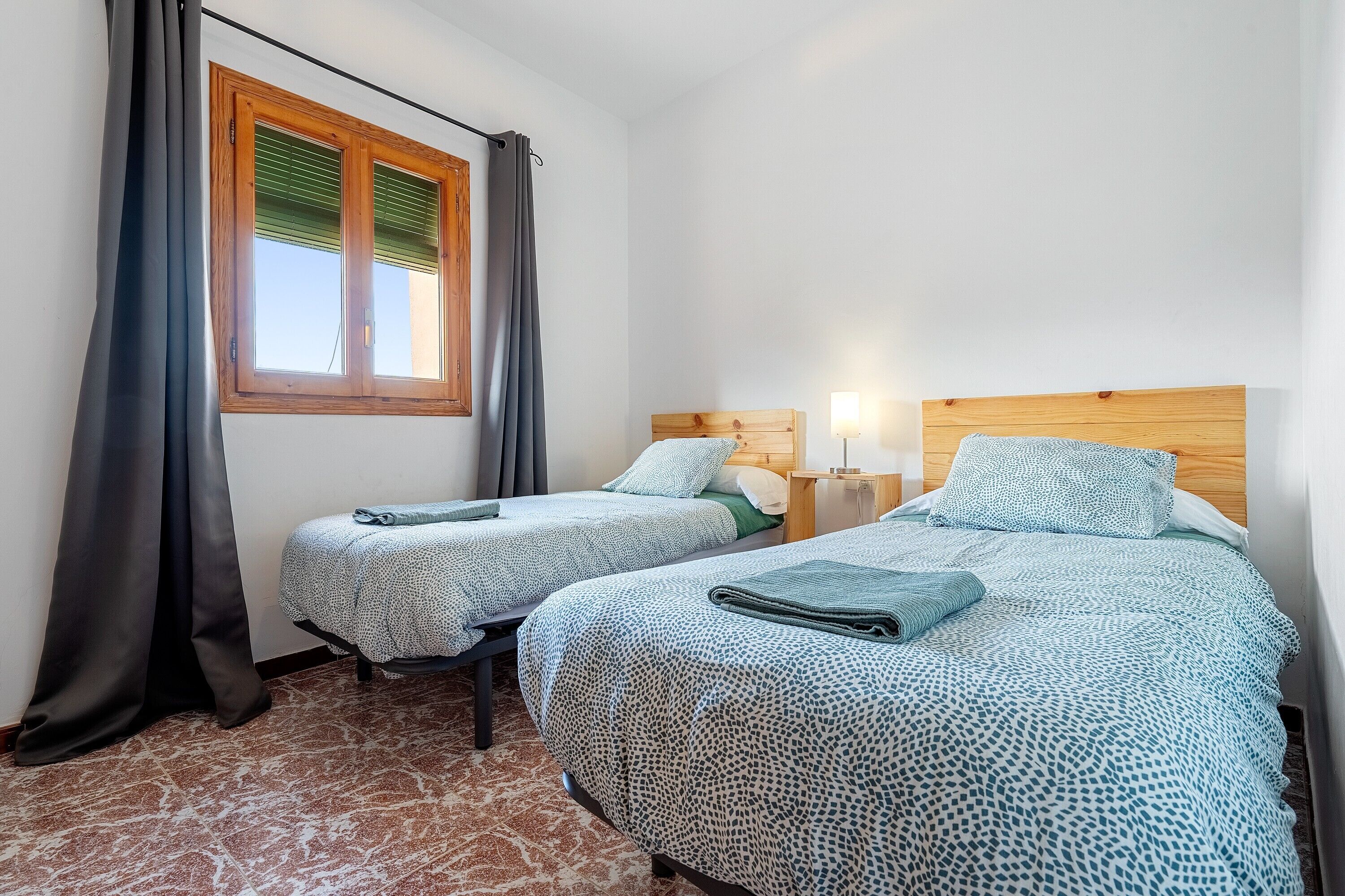 A room with two beds, a window with blinds, a curtain, and a patterned floor.