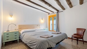 Un dormitorio con cama, mesita de noche, silla y ventana con cortinas.