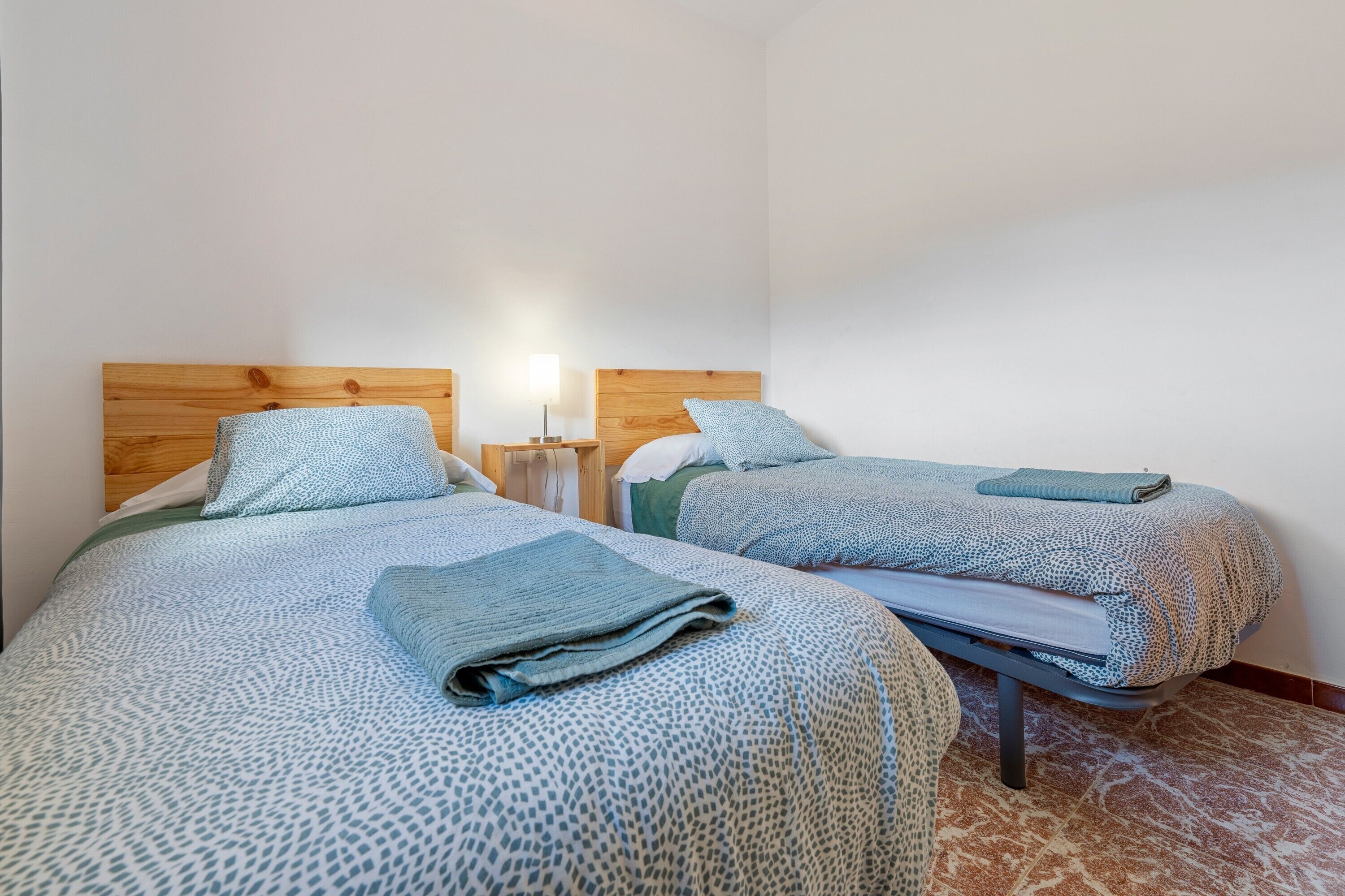 Dos camas con ropa de cama estampada, cabeceras de madera, mesitas de noche con lámparas y suelo de baldosas.