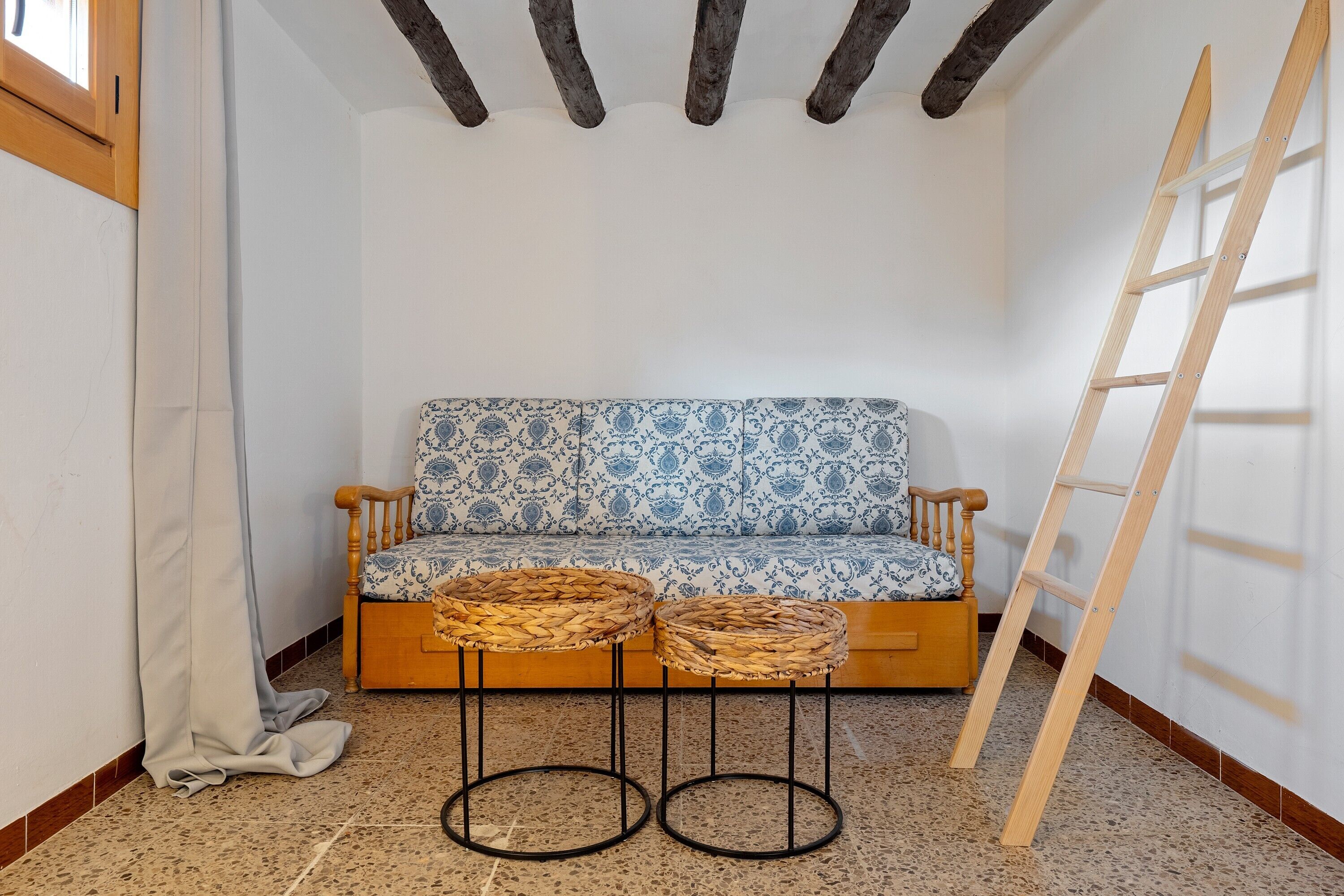 Une pièce d’angle avec un banc en bois, deux tabourets en osier, une échelle et un canapé à motifs.