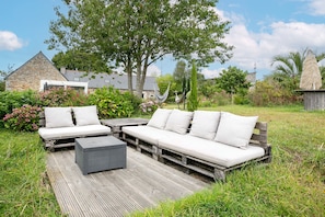 Une terrasse en bois avec un canapé et un pouf, entourée d’une végétation luxuriante et d’un bâtiment en pierre.