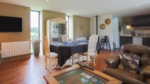 A modern living room with a dining table, leather sofa, and a kitchen in the background. - Maison "Keryanti" with Jacuzzi, Private Terrace, Garden & Wi-Fi (Plouégat-Moysan)