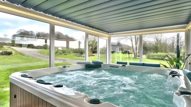 A hot tub under a covered patio with a view of a grassy area and a bridge. - Maison "Keryanti" with Jacuzzi, Private Terrace, Garden & Wi-Fi (Plouégat-Moysan)
