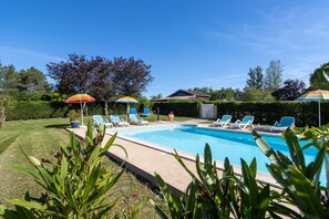 Piscina con sillas de descanso y sombrillas, rodeada de follaje y un cielo despejado.