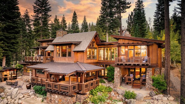A large wooden cabin with multiple decks and outdoor seating areas, surrounded by a rocky landscape and dense forest.