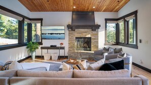 A spacious living room with a stone fireplace, large windows, and a wooden ceiling.