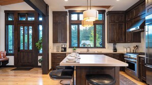 A modern kitchen with dark wood cabinets, a large island with bar stools, a stainless steel range hood, and large windows offering a view of the outdoors.