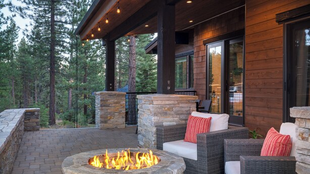 A stone fire pit surrounded by wicker furniture with cushions, set on a paved patio with a view of a forested area.