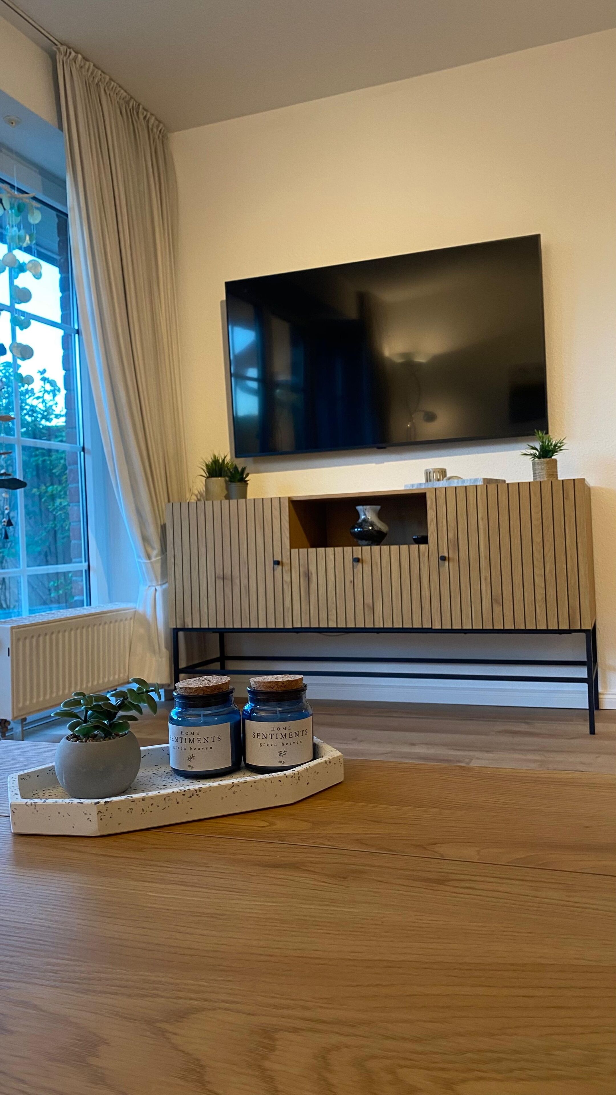 Ein modernes Wohnzimmer mit einem großen Flachbildfernseher, der an einer Holzwand angebracht ist, einer Topfpflanze und einer Schale mit zwei Gläsern, beschriftet mit 'HOME SENTIMENTS'.