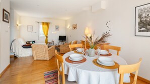 A dining area with a round table set for four, surrounded by wooden chairs, a television in the background, and a cozy seating area with a sofa and cushions.