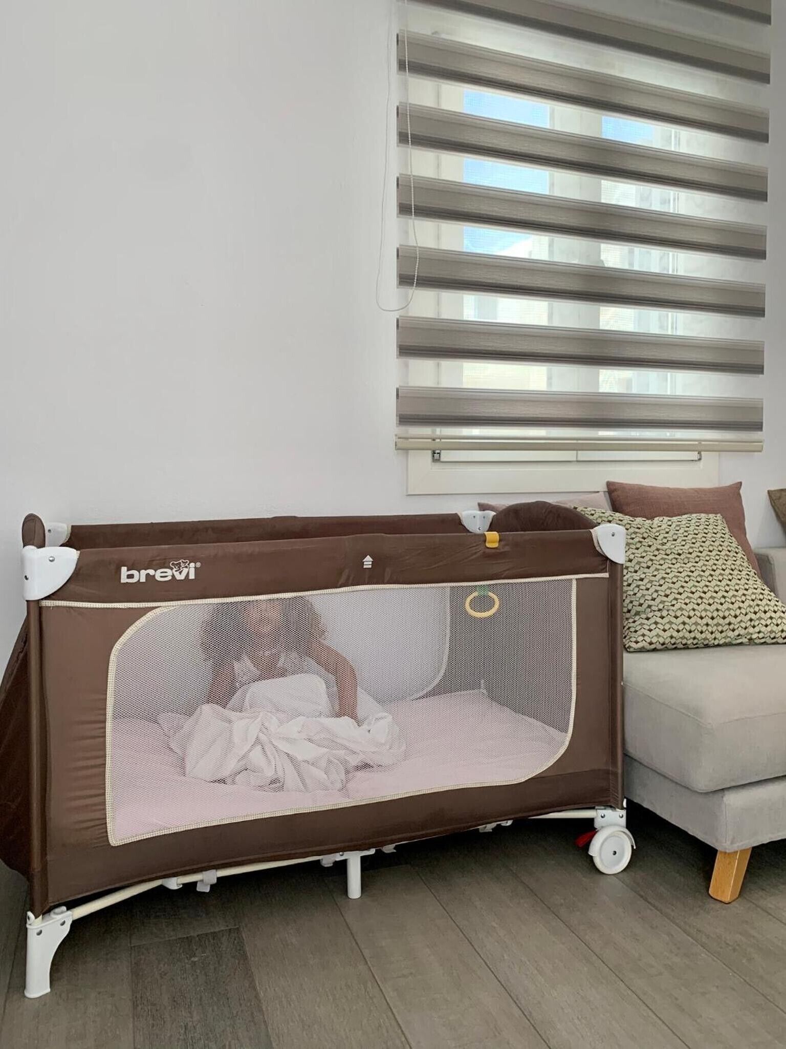Una habitación con una cuna para bebé, un sofá gris y una ventana con persianas.