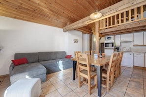 A cozy living area with a wooden ceiling, a grey sofa with red cushions, a dining table with wooden chairs, and a kitchen with white cabinets and appliances. - Apartment 'Le Génépi' with Mountain View, Balcony and Wi-Fi (Val-Cenis)
