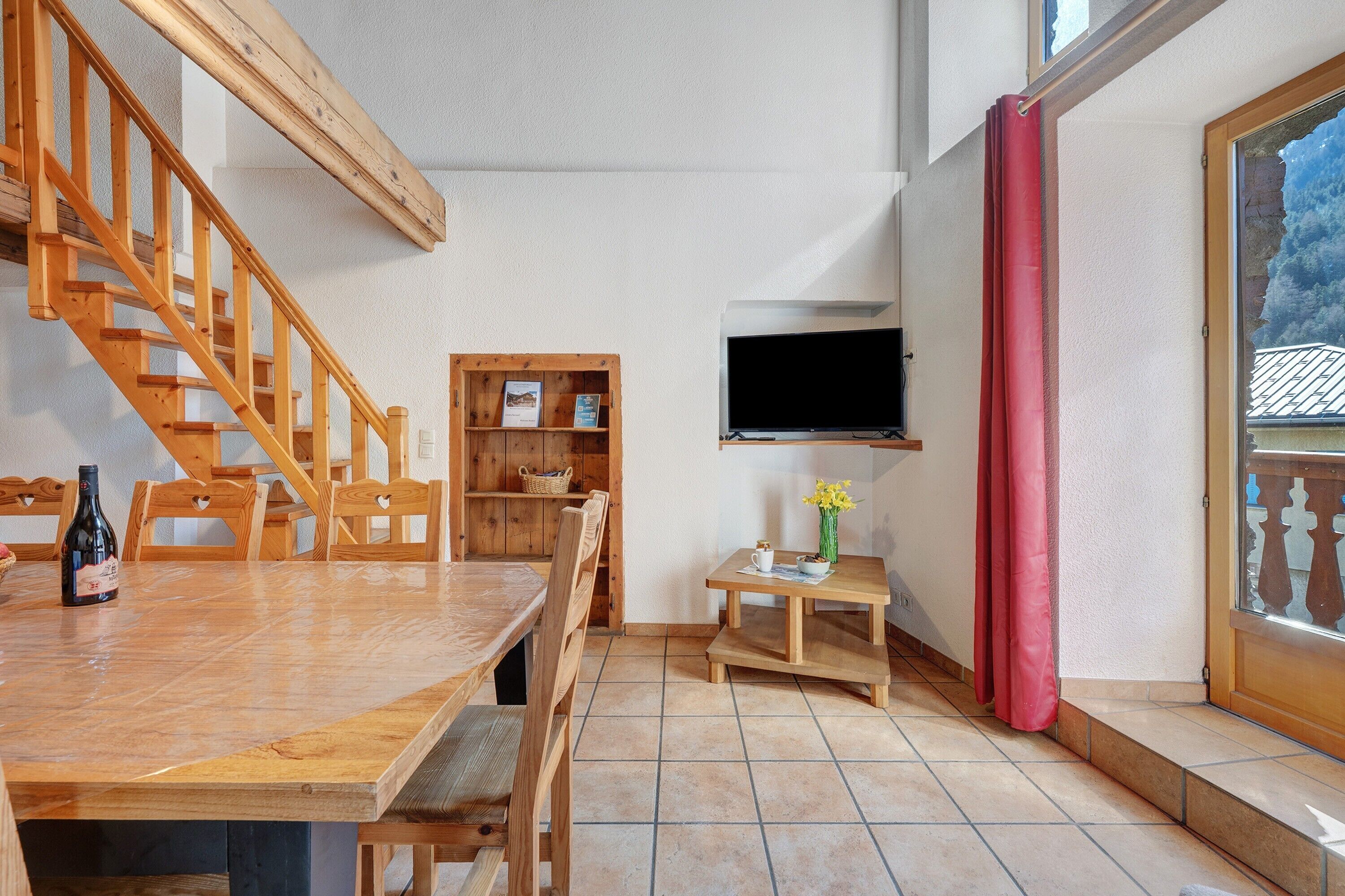 A wooden staircase, a dining table with chairs, a small wooden table with a vase, and a television mounted on the wall.