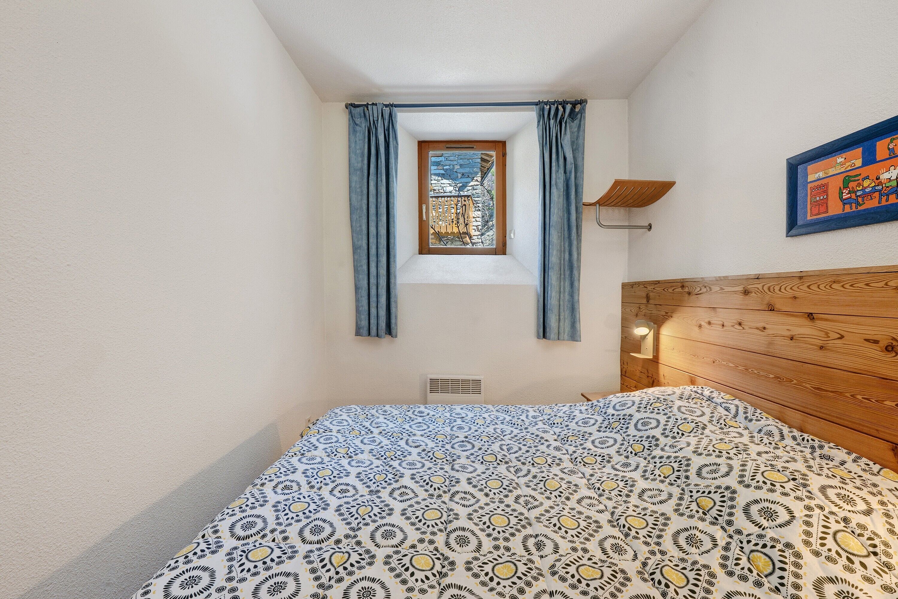 A bedroom with a bed, a window with curtains, a headboard, a wall-mounted light, and a painting.