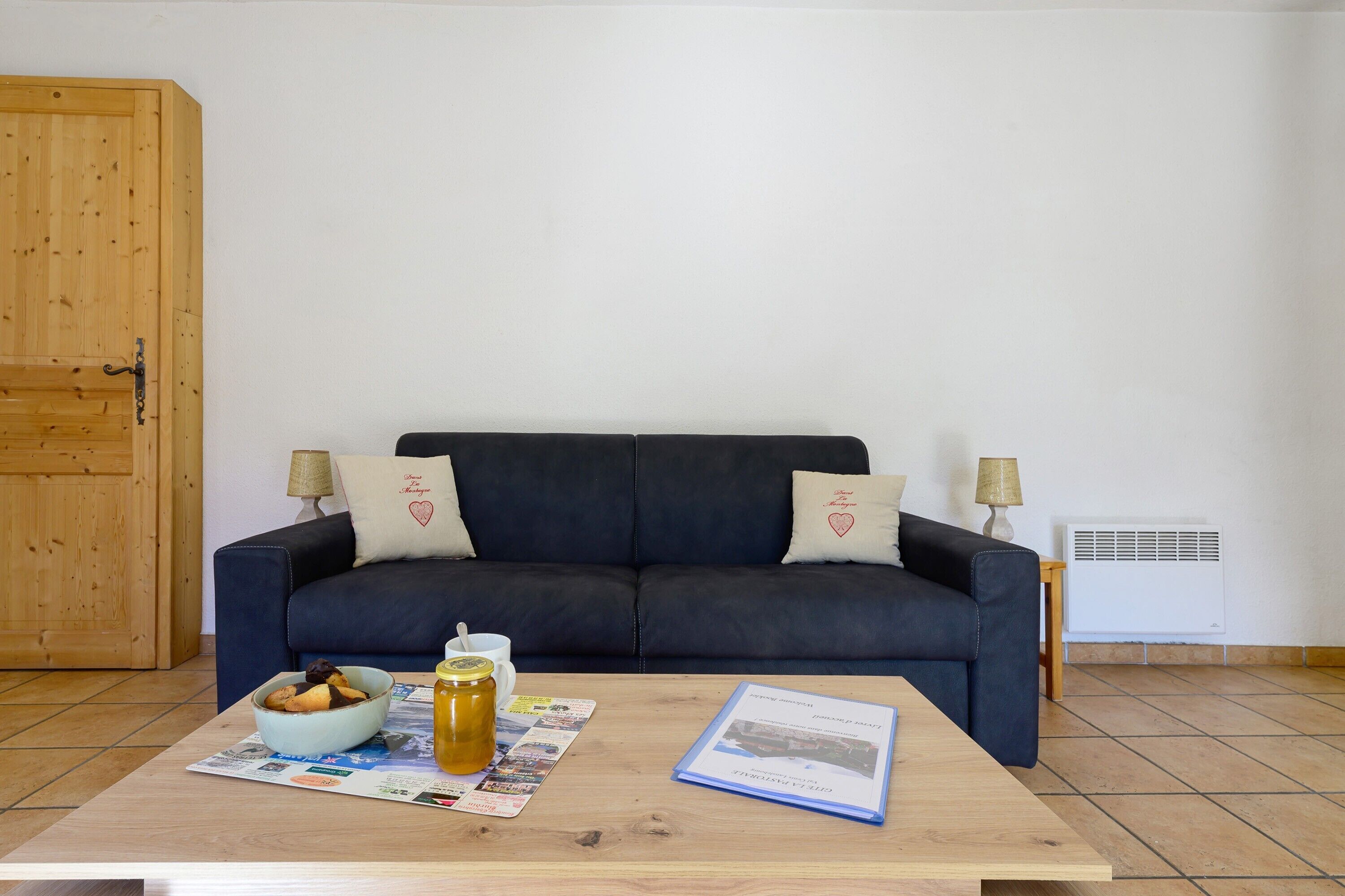 A living room with a dark blue sofa, two lamps, a wooden table with a book, a bowl of cookies, and a jar of honey.