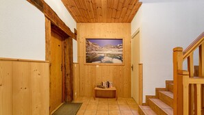 A wooden interior hallway with a wooden door, a framed picture on the wall, a small wooden bench with books, and a staircase. - Apartment 'Les Gentianes' with Mountain View and Wi-Fi (Val-Cenis)