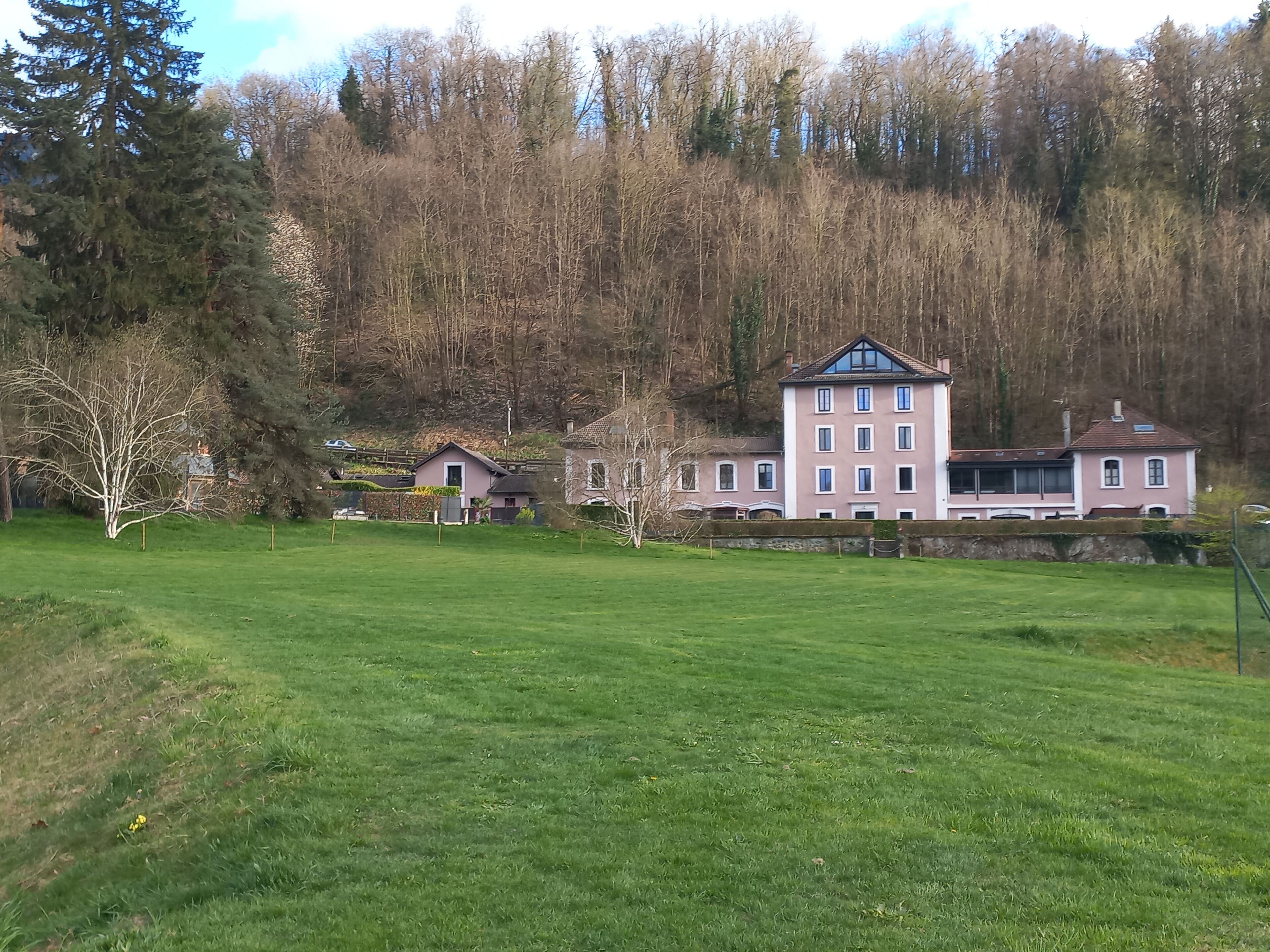 A large building with multiple windows and a prominent entrance, surrounded by a grassy field and dense trees.