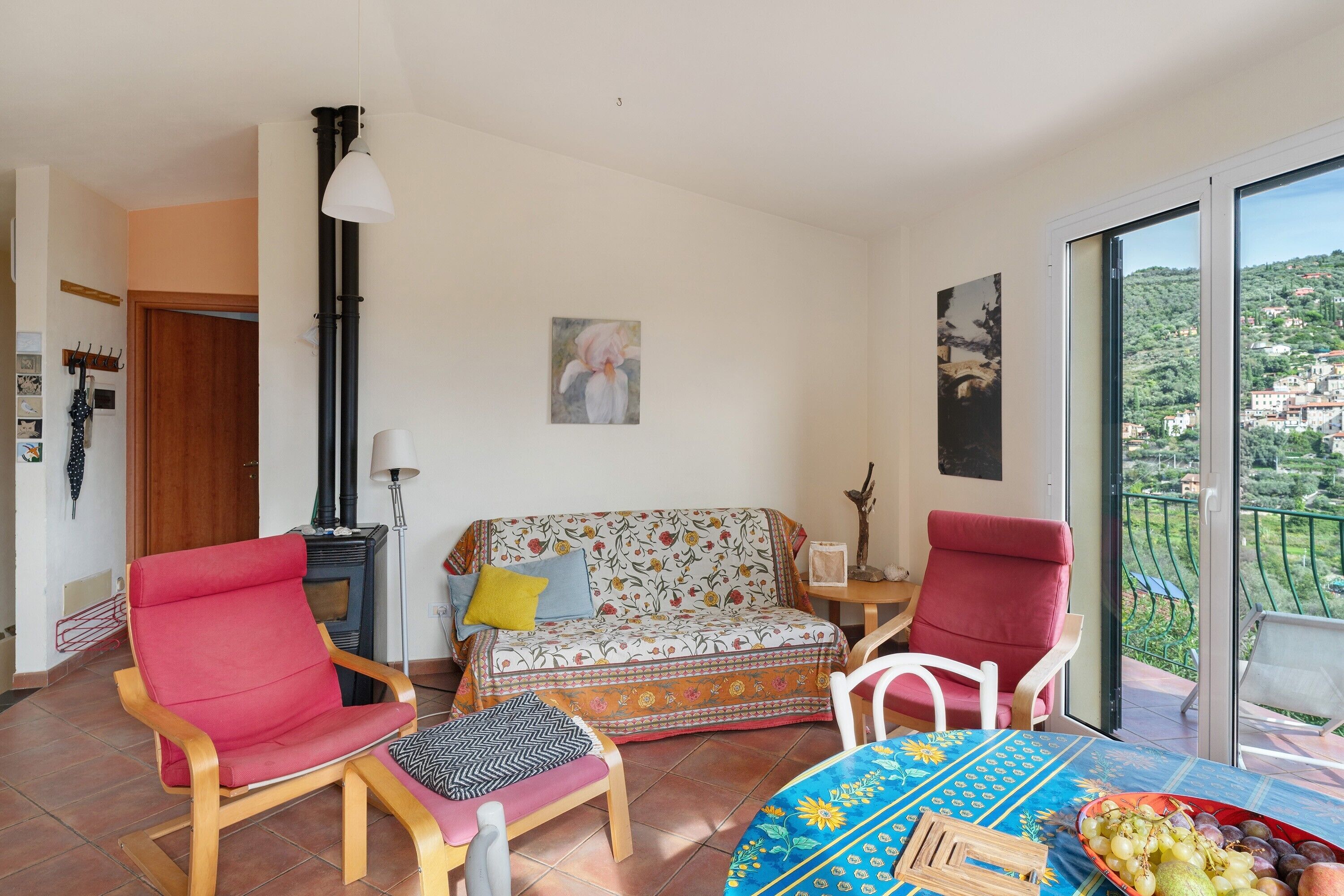 A living room with a sofa, two chairs, a table with fruit, and a view of a hillside.