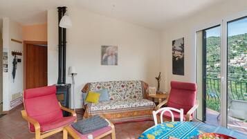 A living room with a sofa, two chairs, a table with fruit, and a view of a hillside.