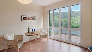 A room with a wicker chair, a small table with a vase and two glasses, and a large window with a view of greenery. - Holiday Home 'Casa Bilger' with Sea View, Private Terrace and Wi-Fi (Dolcedo)