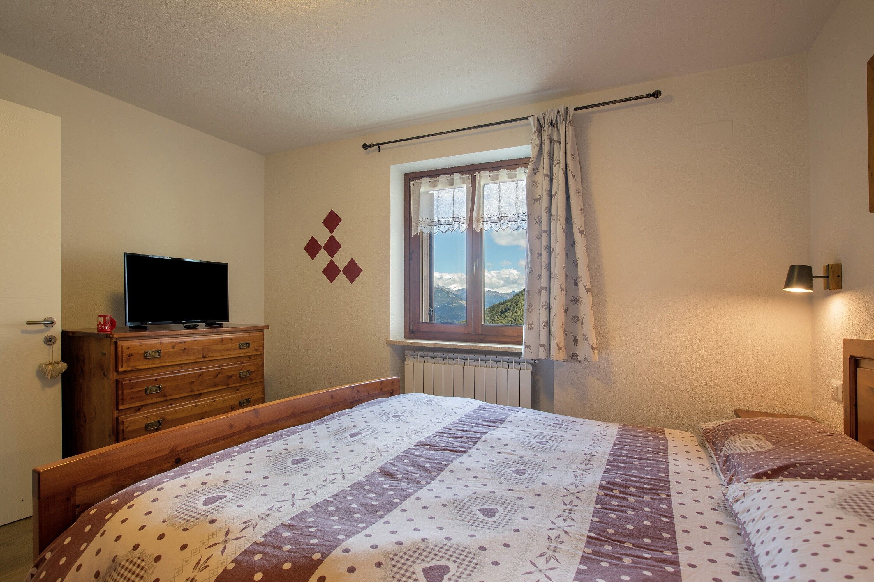 Un dormitorio con cama, cómoda, televisor encima y ventana con cortinas, y vistas a las montañas.