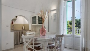 A dining area with a white table and chairs, a vase with dried plants, and a view through a glass door to the outside.