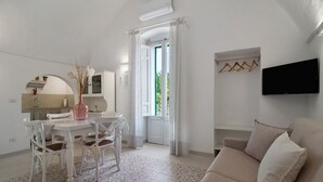 A modern living room with a sofa, a dining table with chairs, a TV mounted on the wall, and a kitchen area visible through an open door.