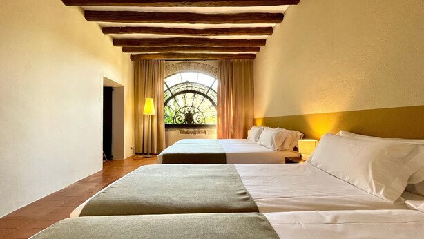A hotel room with two beds, a window with curtains, a lamp, and a wooden ceiling. - Apartment 'Apartaments Can Gibert - Muralla' with Shared Garden, Balcony and Wi-Fi (Castelló d'Empúries)