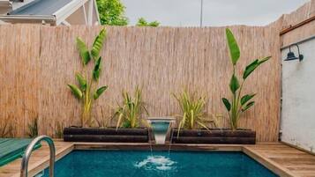 Ein rechteckiger Pool mit einer Holzterrasse, umgeben von einem Bambuszaun und verschiedenen Pflanzen.