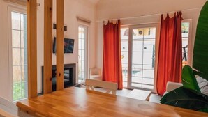 Une pièce avec une table à manger en bois, des chaises blanches, un téléviseur fixé au mur et une fenêtre ornée de rideaux rouges.